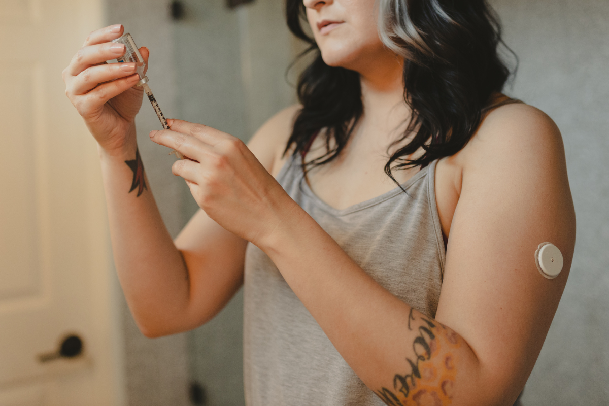 A young white woman is photographed filling a syringe, she has a blood sugar monitor on her arm. 