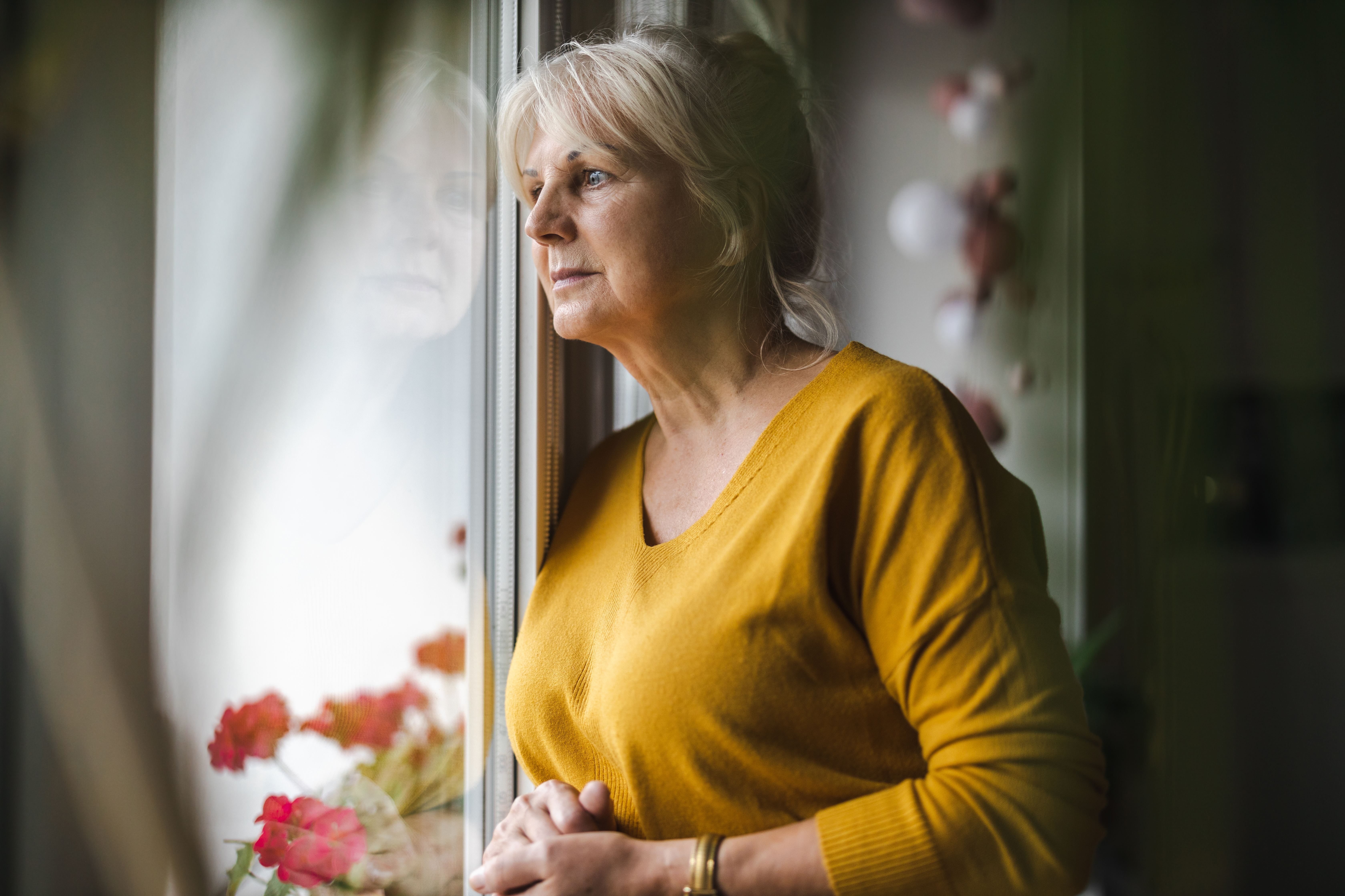 An older woman wearing a yellow jumper looking out of a window.