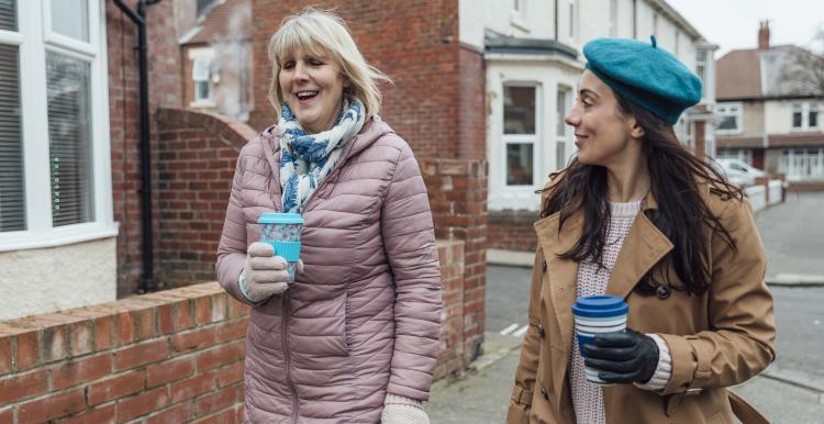 Two women drinking hot drinks out of reusable cups walking down a street.