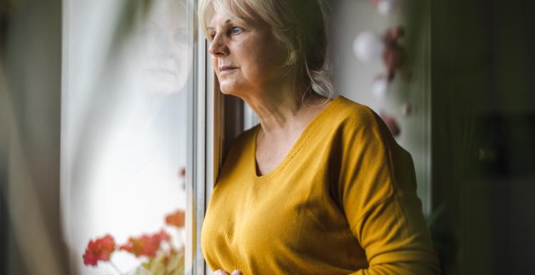 An older woman wearing a yellow jumper looking out of a window.