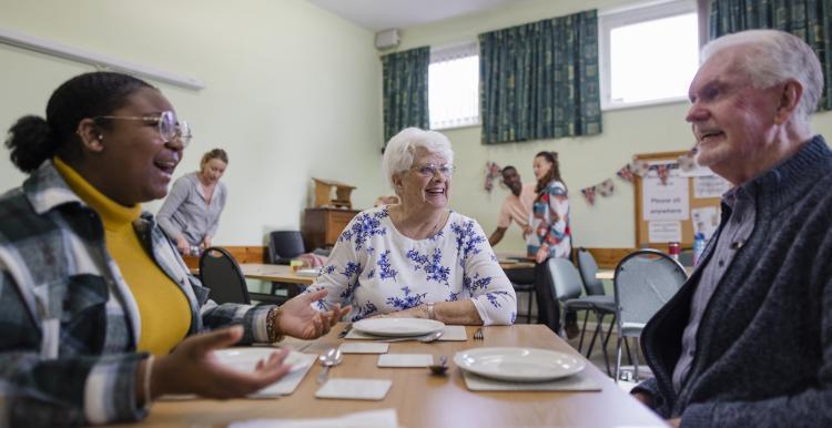 A community church hall, running a soup kitchen and warm hub where three people are chatting sat at a table.