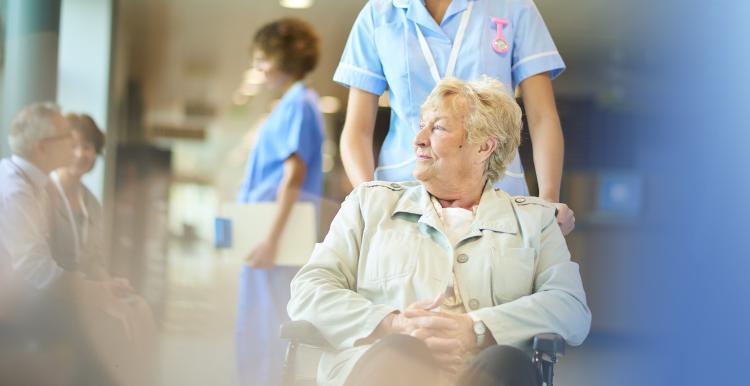 Female patient being pushed in wheelchair through ward by a nurse.