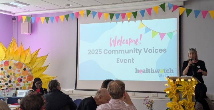 Image shows attendees sat down at tables facing a projector displaying the following message 'Welcome! 2025 Community Voices Event'. The Healthwatch Chief Officer is stood next to the projector on a stand and giving a speech.
