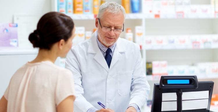 A pharmacist writes a prescription behind a counter while a customer stands nearby
