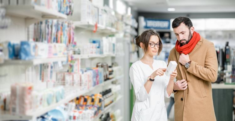 A pharmacist standing in a pharmacy isle with a customer pointing out information on a medication bottle. 