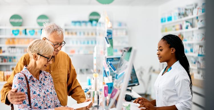 An older couple stand at the counter of a pharmacy and are speaking with the pharmacist. 