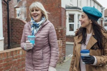 Two women drinking hot drinks out of reusable cups walking down a street.