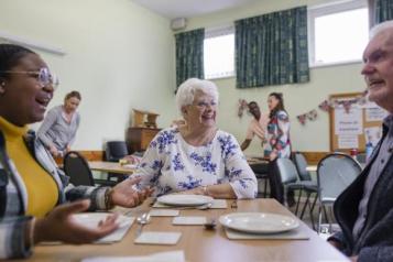 A community church hall, running a soup kitchen and warm hub where three people are chatting sat at a table.