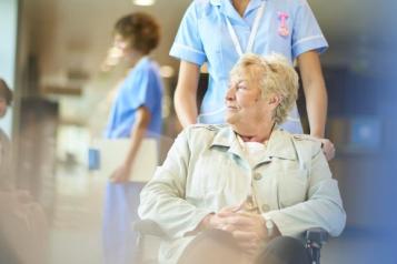 Female patient being pushed in wheelchair through ward by a nurse.