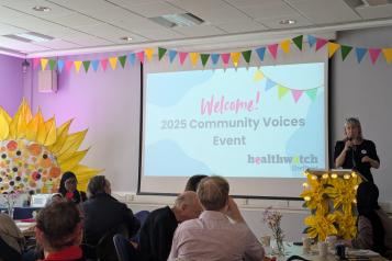 Image shows attendees sat down at tables facing a projector displaying the following message 'Welcome! 2025 Community Voices Event'. The Healthwatch Chief Officer is stood next to the projector on a stand and giving a speech.
