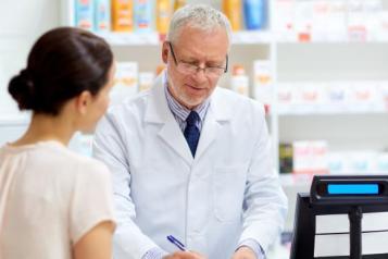 A pharmacist writes a prescription behind a counter while a customer stands nearby