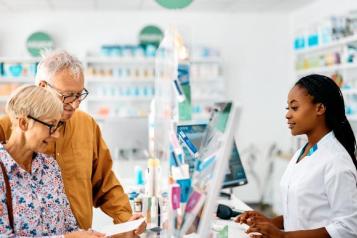 An older couple stand at the counter of a pharmacy and are speaking with the pharmacist. 